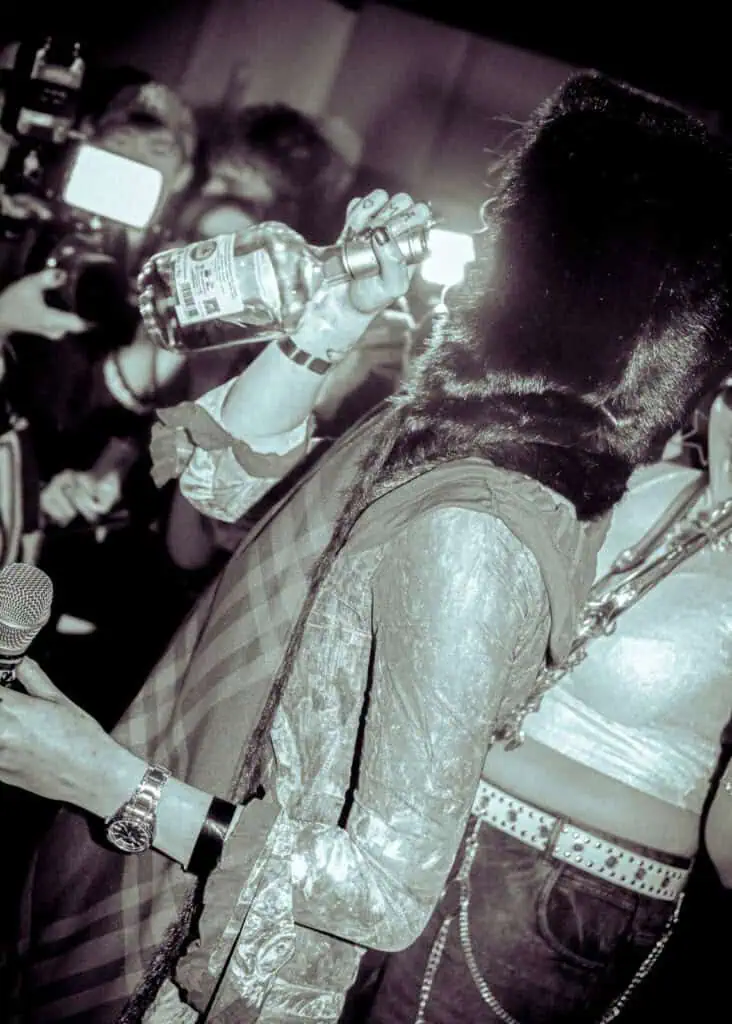 Person wearing a furry hat drinks from a liquor bottle at a crowded party, holding a microphone. Flashing cameras and other people are visible in the background. The image has a vintage, high-contrast effect.