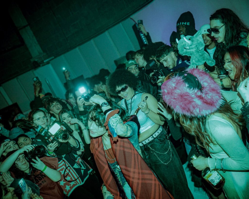 A lively crowd at an indoor event, with people dancing, cheering, taking photos, and flashing cameras. The lighting is dim and colorful, creating an energetic and festive atmosphere.