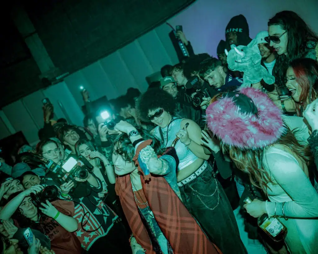 A lively crowd at an indoor event, with people dancing, cheering, taking photos, and flashing cameras. The lighting is dim and colorful, creating an energetic and festive atmosphere.