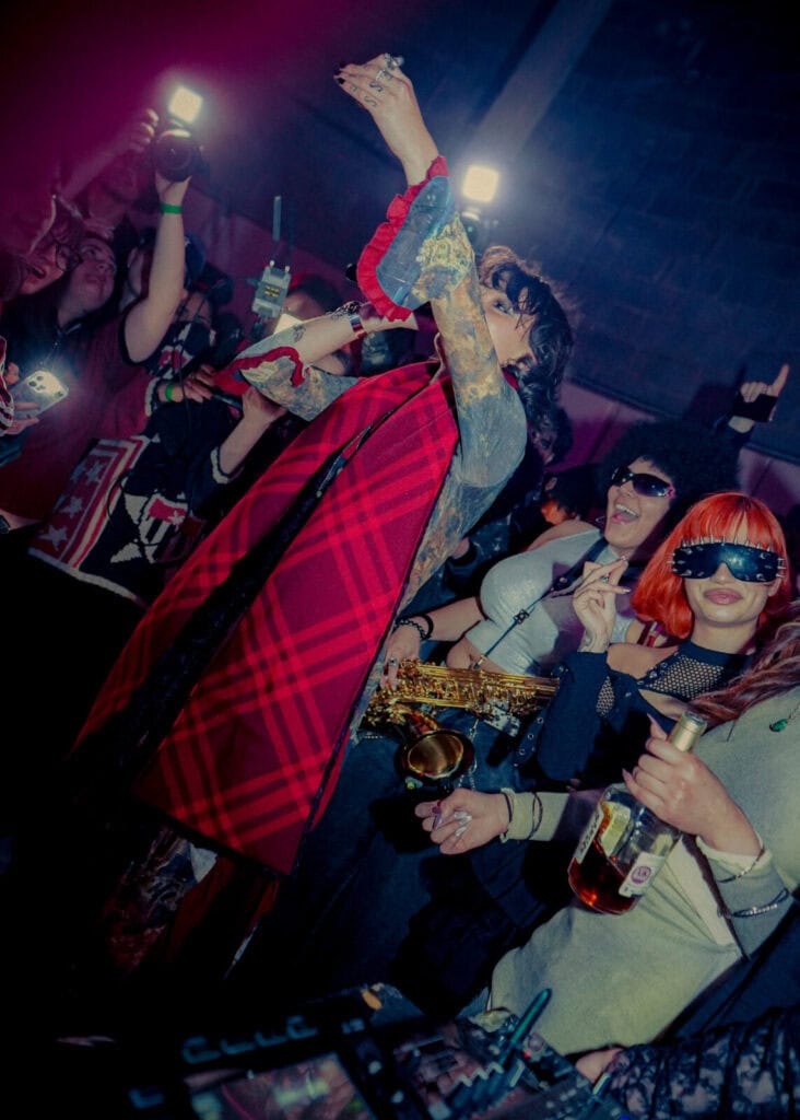 A lively nightclub scene with people dancing and celebrating. One person in the foreground wears a red plaid cape and raises their arm; another holds a saxophone. Others hold drinks and wear sunglasses, smiling under colorful lights.