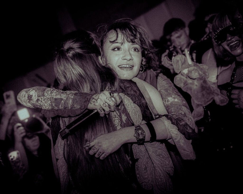 Two people hug tightly on stage during a live performance, one holding a microphone. The crowd in the background cheers and takes photos, some holding stuffed animals. The atmosphere is emotional and energetic.