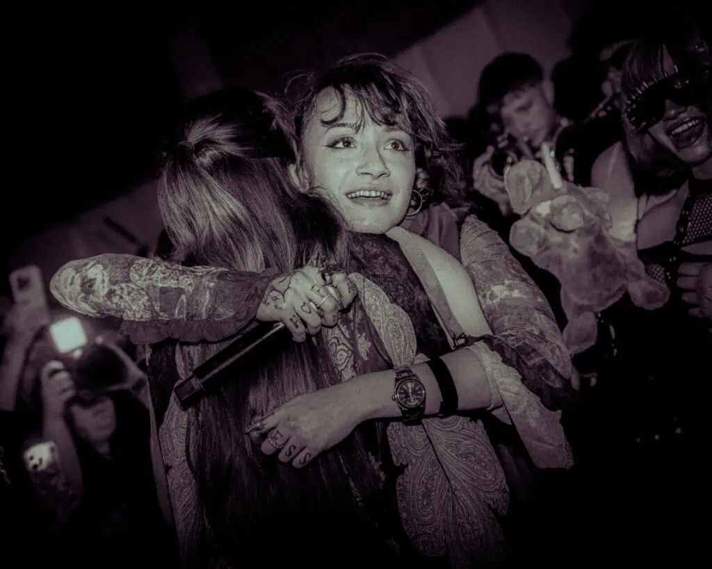 Two people hug tightly on stage during a live performance, one holding a microphone. The crowd in the background cheers and takes photos, some holding stuffed animals. The atmosphere is emotional and energetic.