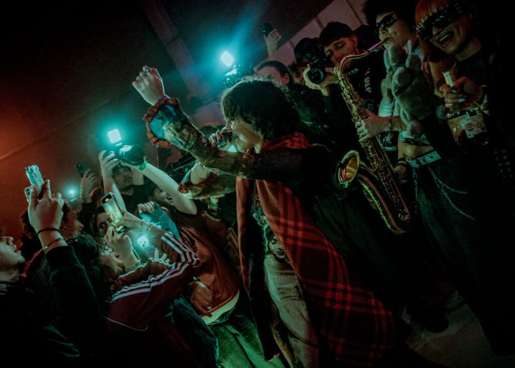 A singer performs passionately in front of a lively crowd, many holding up phones and cameras. Musicians with instruments, including a saxophone, join the energetic scene under colorful, dramatic lighting.