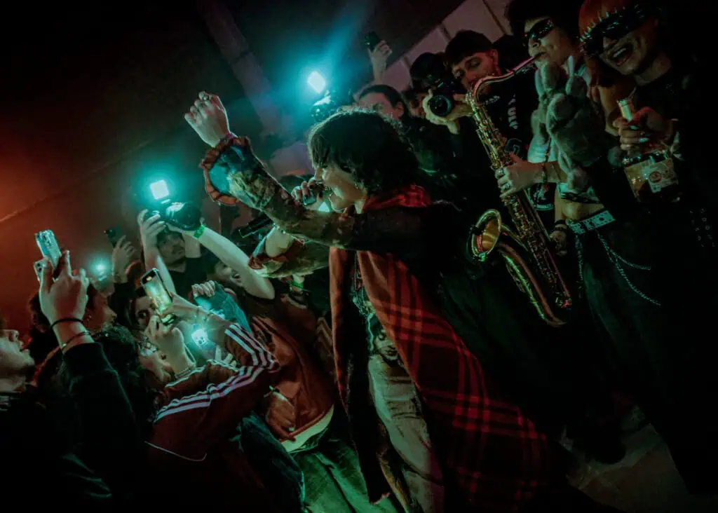 A singer performs passionately in front of a lively crowd, many holding up phones and cameras. Musicians with instruments, including a saxophone, join the energetic scene under colorful, dramatic lighting.