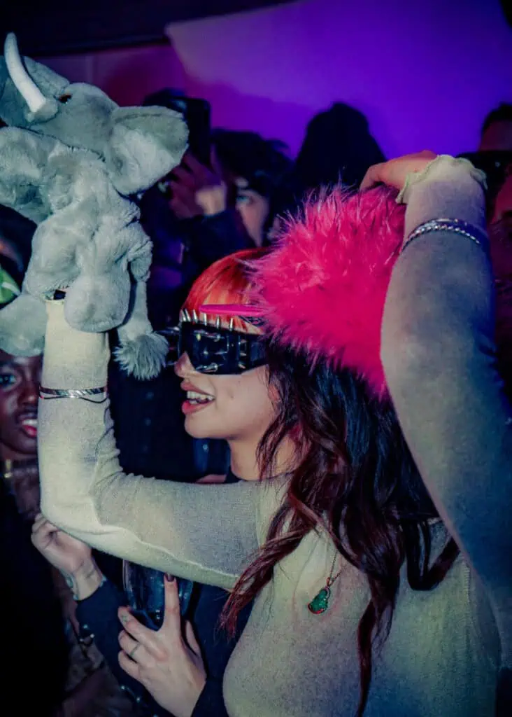 A woman in a sheer long-sleeve top and furry pink hat holds up a plush elephant and smiles. She wears futuristic sunglasses, red-tinted hair, and is surrounded by people in a lively, colorful party scene.
