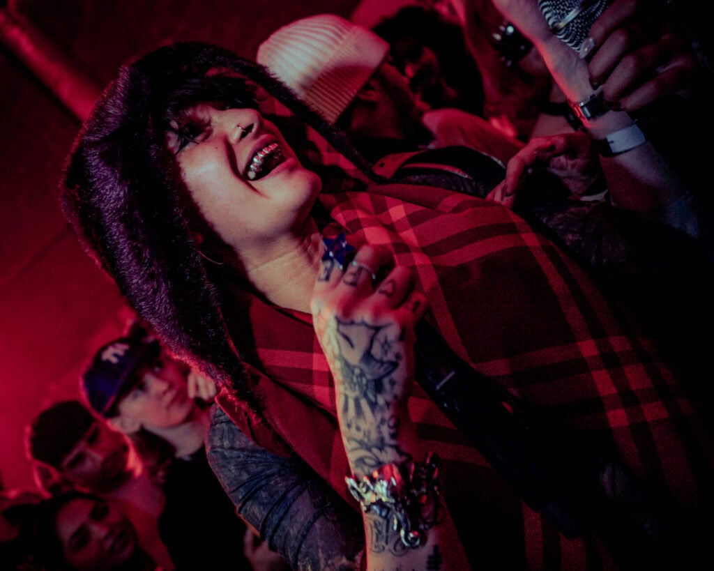 A smiling performer with tattoos and piercings holds a microphone among a crowd, wearing a red plaid scarf and furry hood under pinkish stage lights. Audience members watch closely in the background.