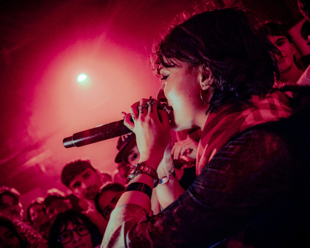 A singer with short dark hair performs passionately on stage, holding a microphone close, while a crowd of people watches in a venue bathed in dramatic red lighting.