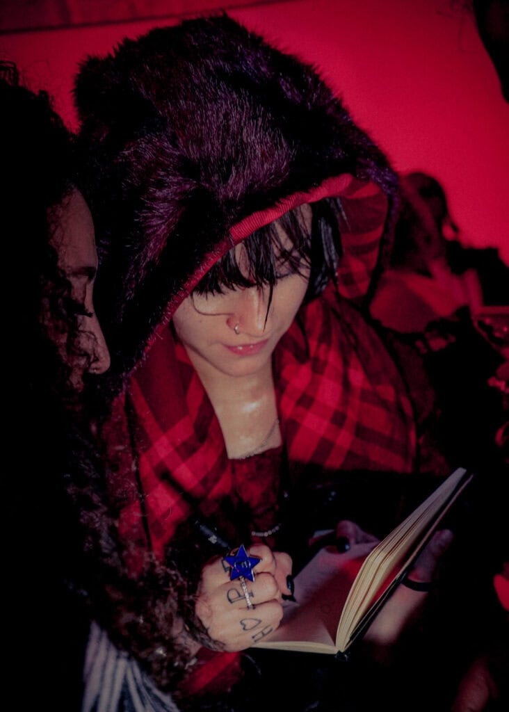A person in a fur-trimmed hood and red plaid scarf signs a notebook. They have a nose ring, dark makeup, and tattoos on their fingers. The scene is lit with a red light, and another person stands close beside them.