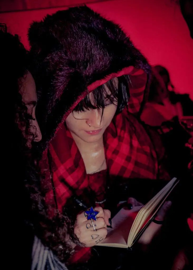 A person in a fur-trimmed hood and red plaid scarf signs a notebook. They have a nose ring, dark makeup, and tattoos on their fingers. The scene is lit with a red light, and another person stands close beside them.