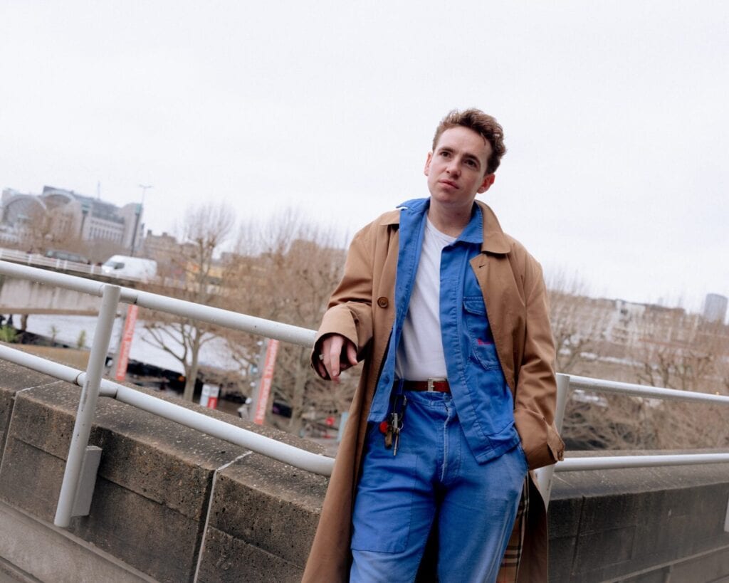 A person stands outdoors, leaning against a railing. They wear a tan trench coat over a blue jumpsuit and white T-shirt. The background shows a cityscape with bare trees and overcast skies.