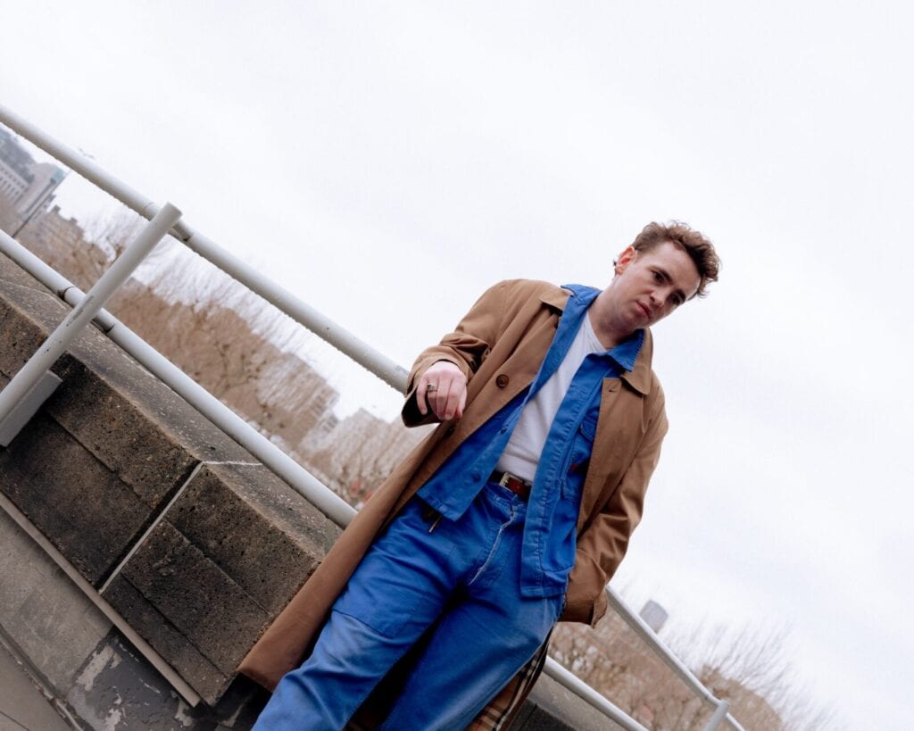 A person stands outdoors by a railing, wearing a brown trench coat over a blue denim jacket and jeans, with a white shirt underneath. The sky is overcast, and city buildings and bare trees are visible in the background.