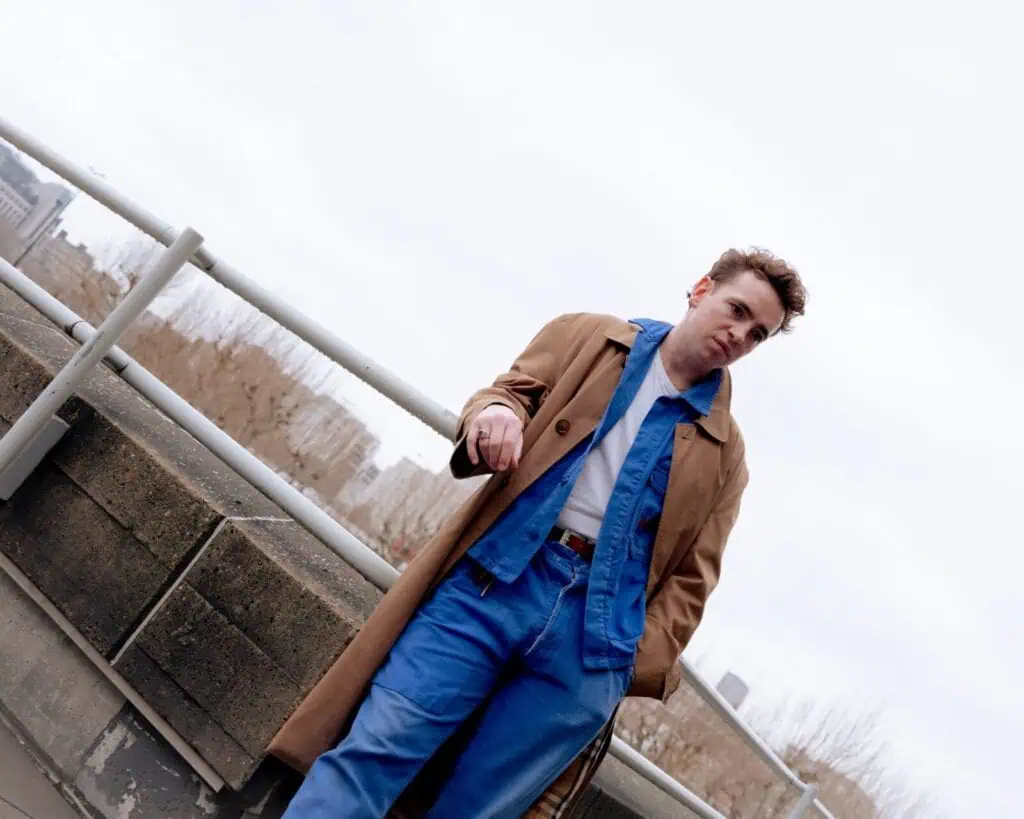 A person stands outdoors by a railing, wearing a brown trench coat over a blue denim jacket and jeans, with a white shirt underneath. The sky is overcast, and city buildings and bare trees are visible in the background.