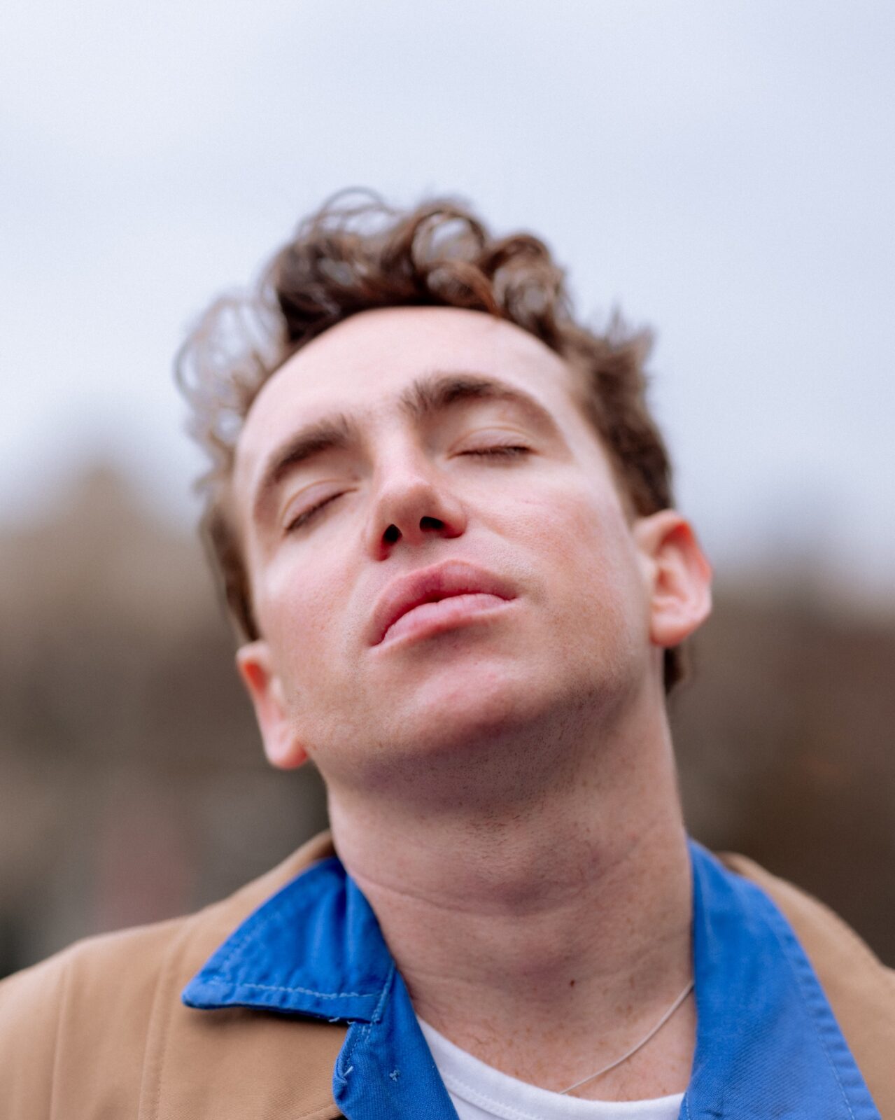 A person with short, curly brown hair and closed eyes tilts their head back slightly, appearing relaxed or contemplative. They wear a blue collared shirt under a tan jacket, with a blurred outdoor background.