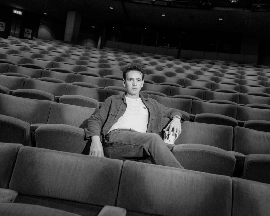 A person sits alone in the middle of an empty theater, leaning back with one arm resting on an adjacent seat and holding a drink container. The rows of unoccupied seats surround them. The image is in black and white.