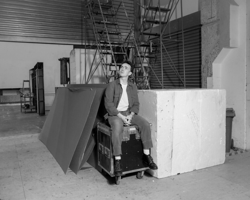 A man in casual clothes sits on a box in an industrial setting, surrounded by large panels and scaffolding. He looks upward thoughtfully. The photo is black and white.