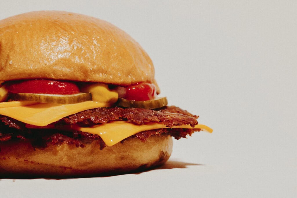 A close-up of a cheeseburger with beef patty, cheddar cheese, pickles, ketchup, and mustard in a soft bun—perfect inspiration if you're searching for where to eat during London Fashion Week. Set against a plain light background.