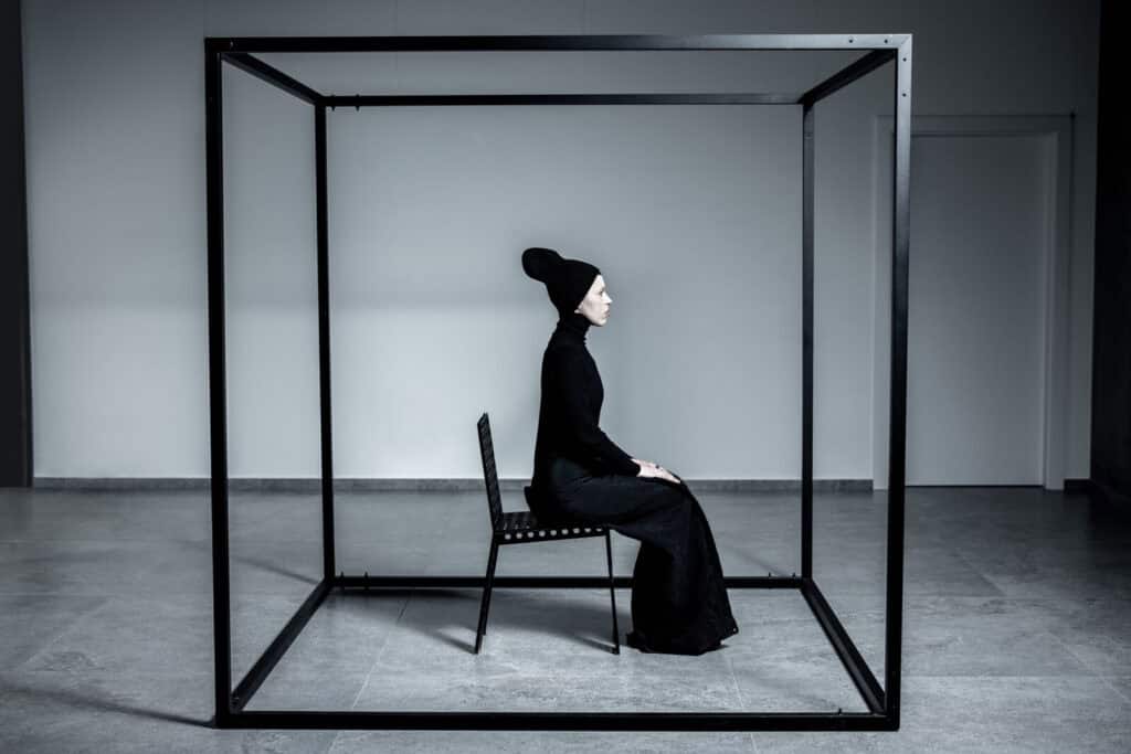 A person dressed in all black sits on a chair inside a large, open black metal cube frame in a minimalist, white room. The person is in profile, wearing a tall hat, and appears contemplative.