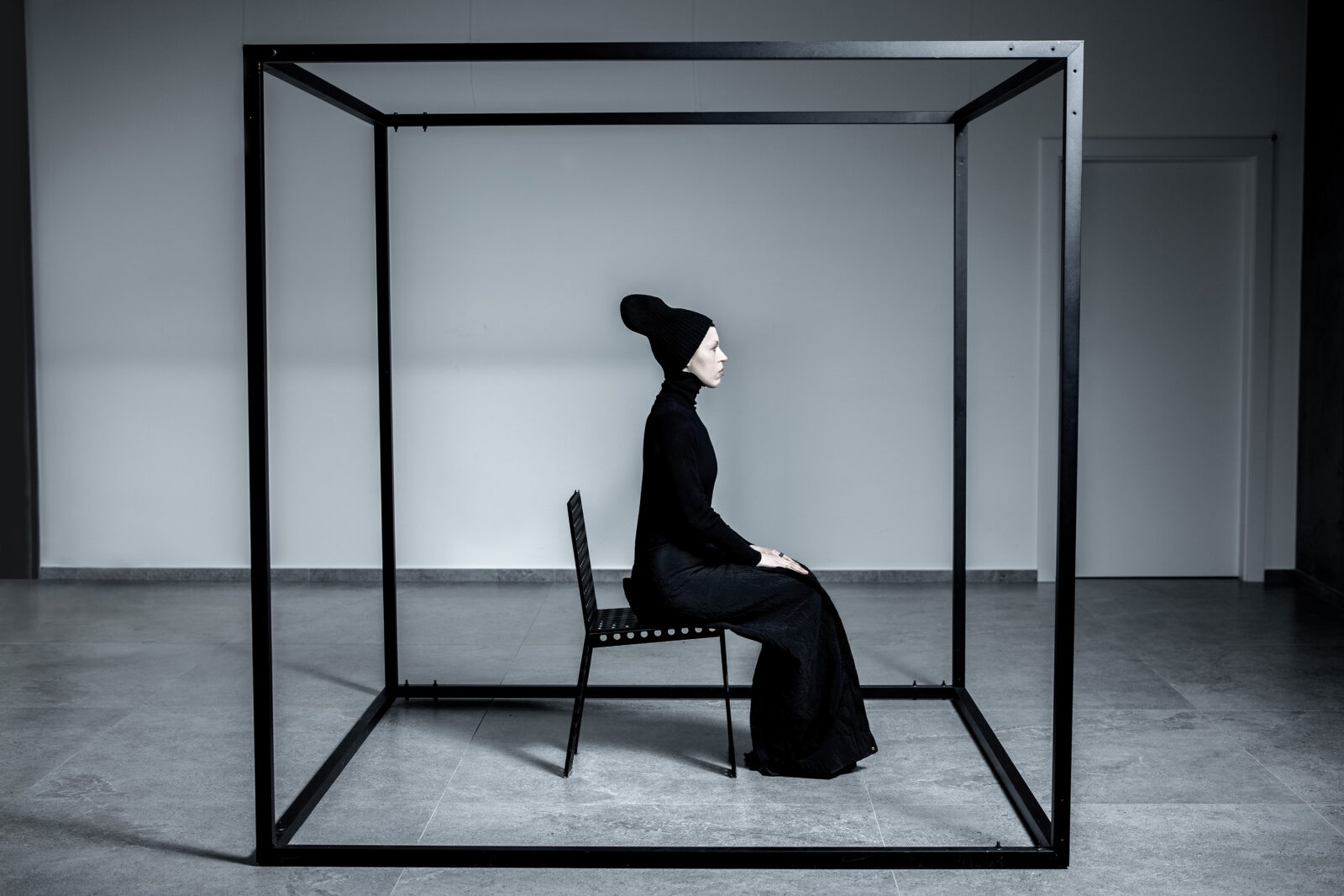 A person dressed in all black sits on a chair inside a large, open black metal cube frame in a minimalist, white room. The person is in profile, wearing a tall hat, and appears contemplative.