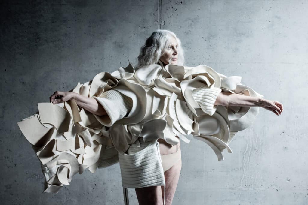 An older woman with long white hair stands against a textured gray wall, wearing an elaborate, sculptural cream-colored outfit with large, layered, ruffled shapes extending from her arms.