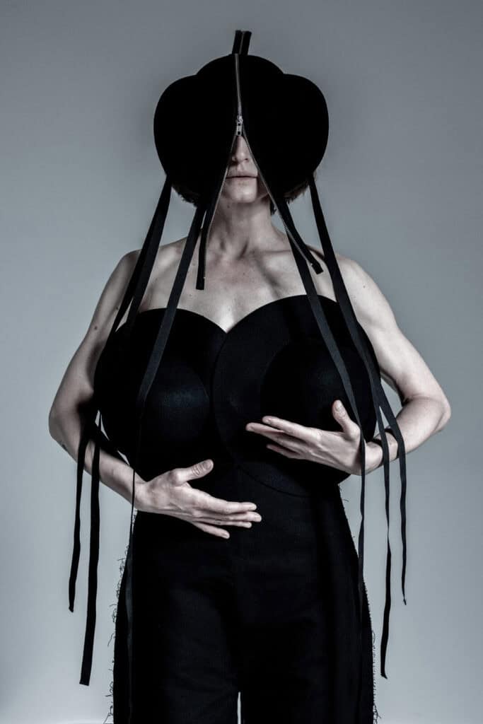 A person in a sculptural black outfit with exaggerated circular shapes on the chest and a headpiece that covers the upper face, featuring long black straps hanging down. The background is plain and light gray.