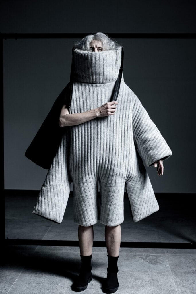 An elderly person stands in a minimalist setting, wearing an oversized, quilted, gray ribbed outfit with wide sleeves and a tall, padded collar covering their neck and mouth, holding a black strap over one shoulder.