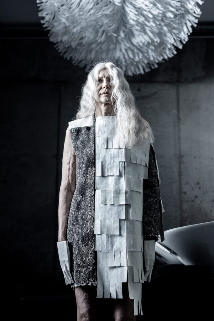 An older person with long white hair stands in a dramatic, futuristic outfit made of black, white, and silver rectangular panels. A large, white, fringed circular art piece hangs overhead against a dark background.