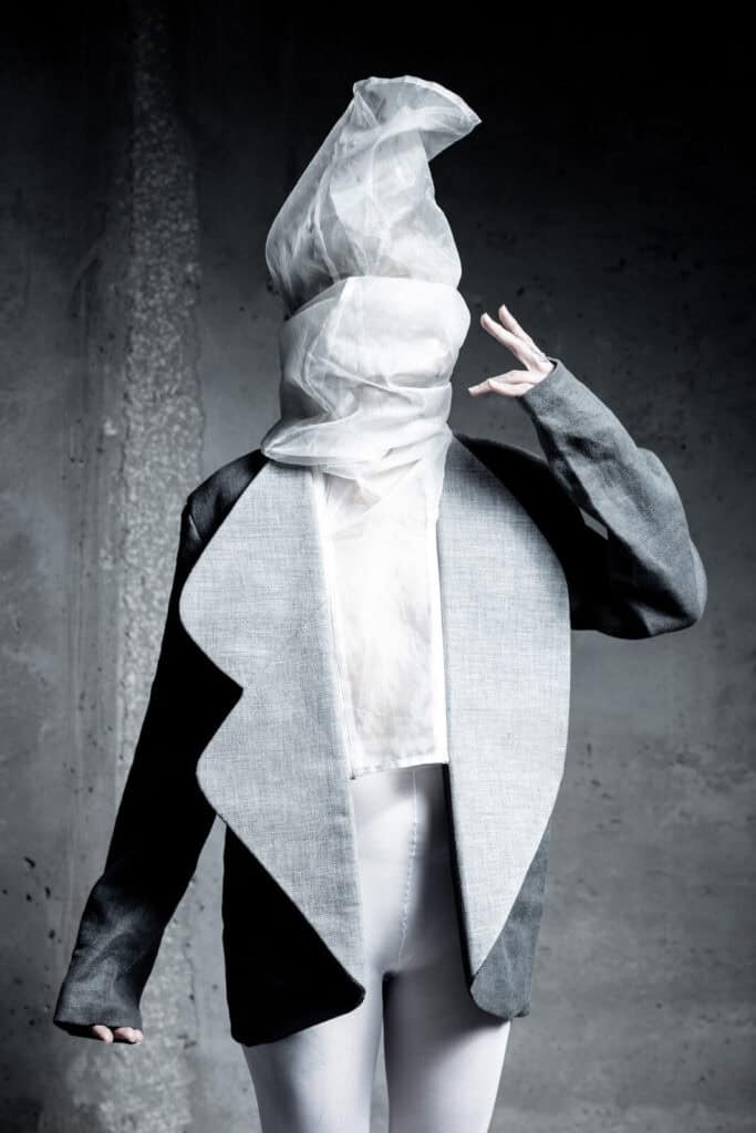 A person wearing an oversized gray and black jacket with white leggings, their head wrapped in sheer white fabric, obscuring their face, posing against a rough concrete wall.