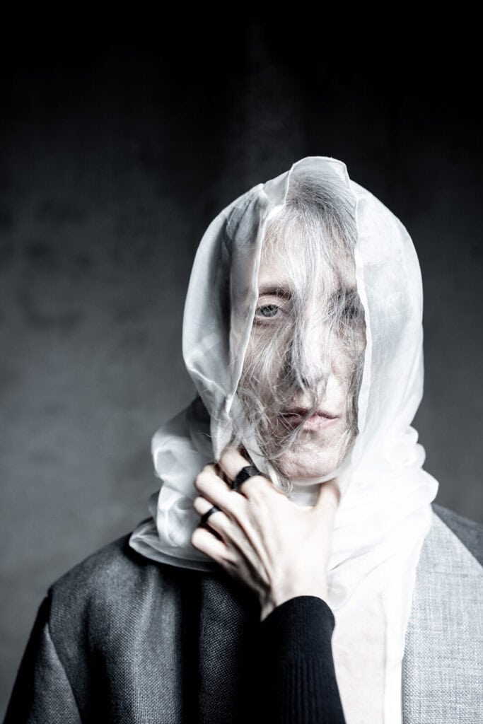 A person wrapped in sheer white fabric gazes forward, with strands of hair visible through the material. A hand, wearing a black ring, gently grasps their neck. The background is dark and blurred.