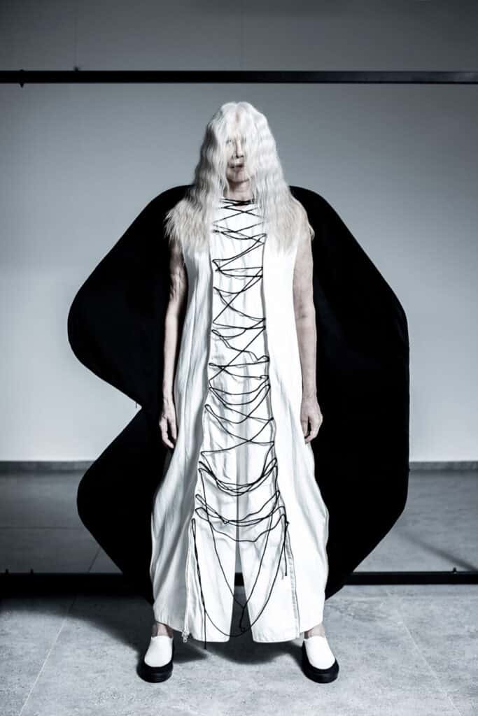 An older person with long white hair stands facing forward, wearing a white dress with black cords crisscrossing down the front and large black sculptural fabric extending from their back, against a plain background.