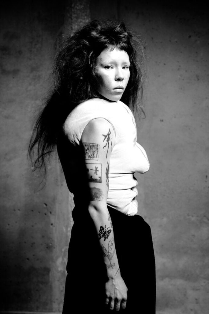 Black and white photo of a person with long, tousled hair wearing a white top and dark pants, looking over their shoulder. Their exposed arm displays several tattoos. The background is a textured wall.