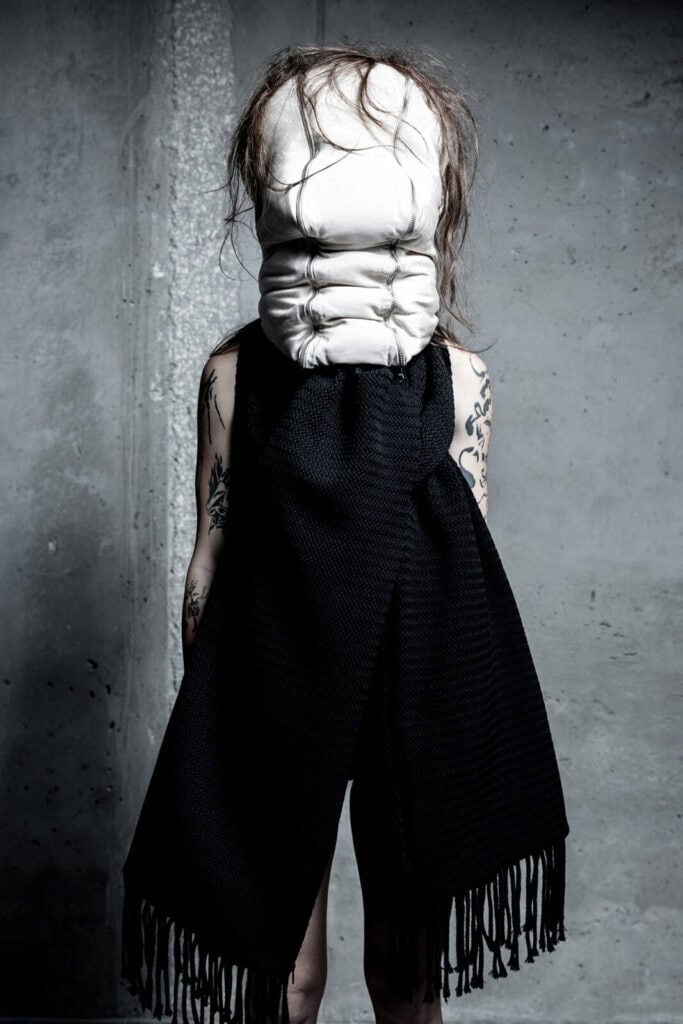 A person with tattooed arms stands against a concrete wall, wearing a large black garment and a white padded mask that obscures their entire head. Their hair is messy and protrudes from the mask.