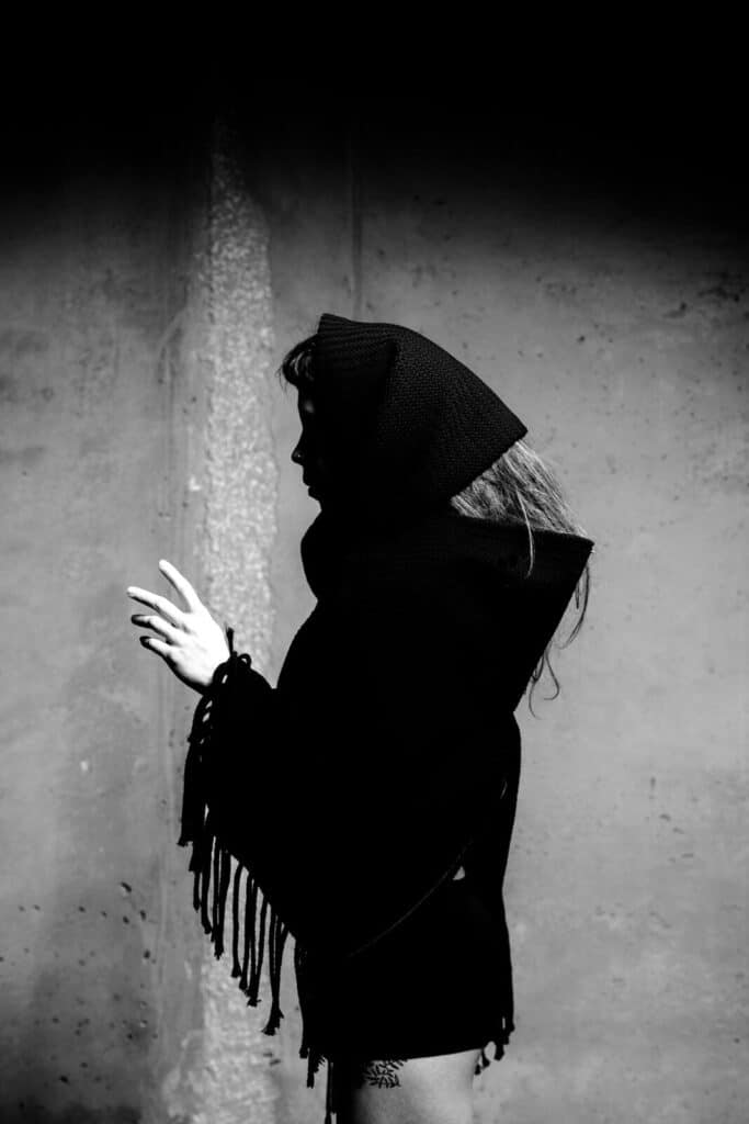 A person in profile wears a dark, hooded garment with fringe, standing against a plain wall. The lighting creates a dramatic shadow, highlighting their silhouette and outstretched hand.