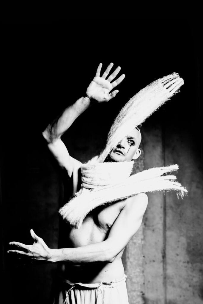 A shirtless person with dramatic makeup poses with arms raised, their face and chest partially obscured by thick, textured white fabric strips, in a high-contrast black and white photo.