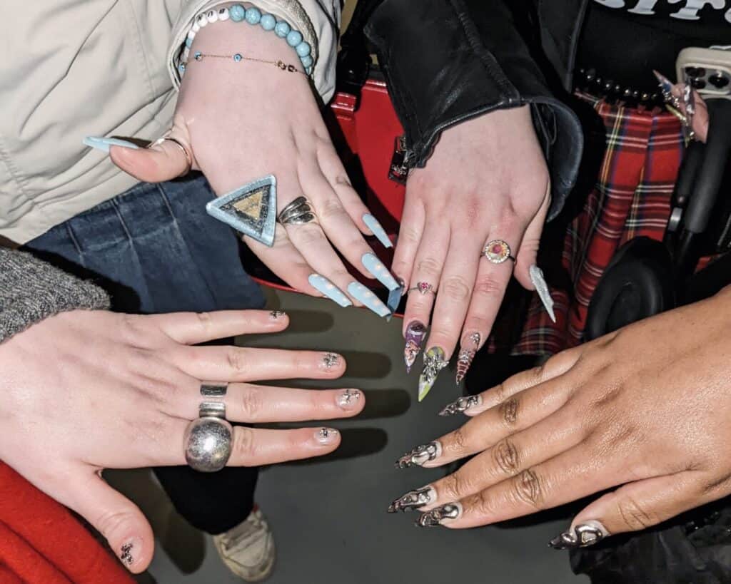 Four hands with long, decorated, and colorful manicured nails—each flaunting unique Valentine-inspired designs and rings—come together above a gray floor. The background shows glimpses of jackets, bracelets, and a red plaid skirt.