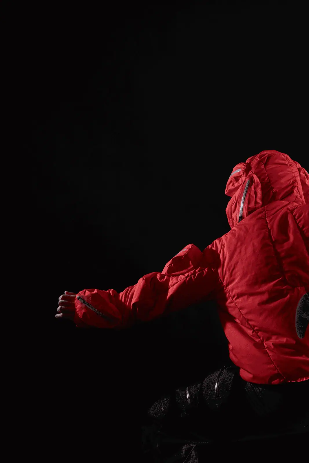 A hooded figure in a bright red jacket sits with their back to the camera against a dark background, extending one arm out to the side.