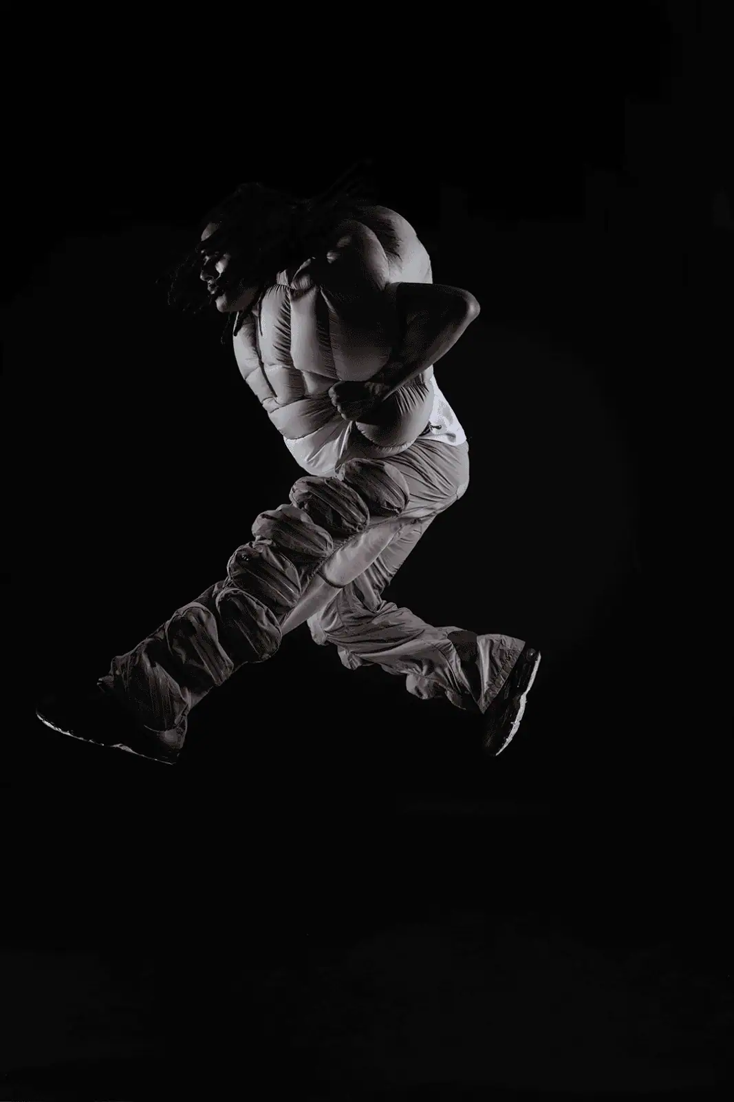 A hooded person with dreadlocks, wearing a white padded vest and baggy gray pants, leaps into the air against a black background. Their body is angled sideways, and their arms are bent in motion.