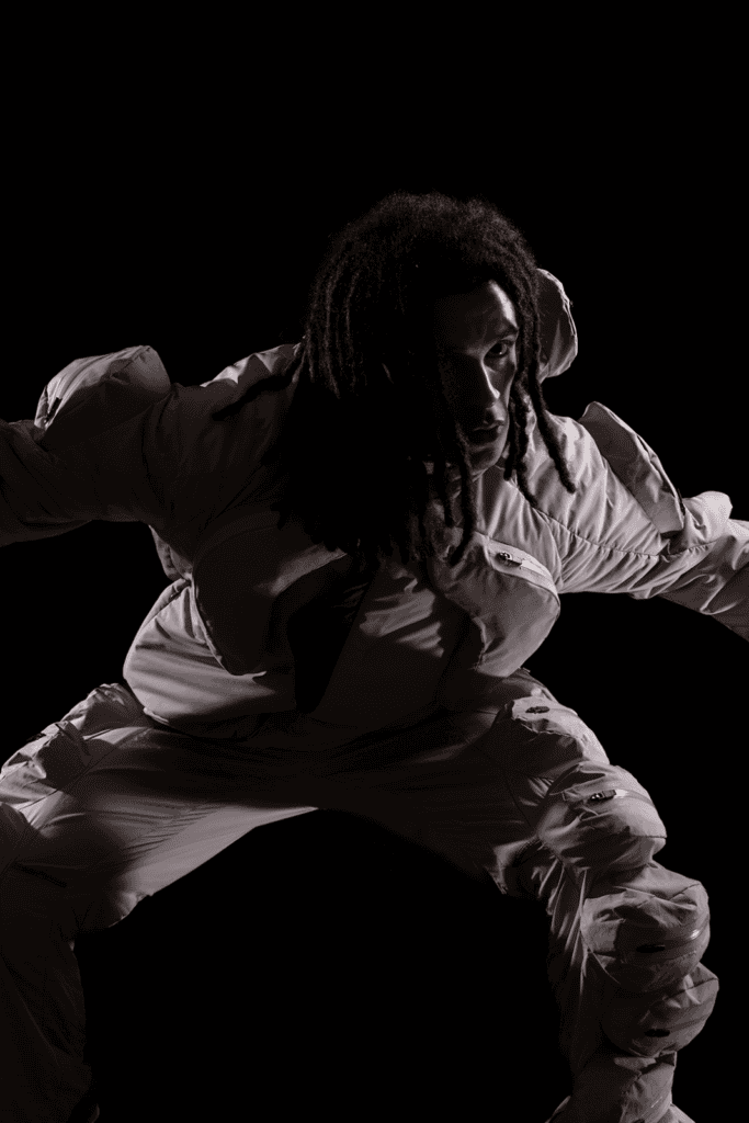 A person with long, dark hair wearing a light-colored, hooded padded suit poses against a dark background, partially crouched with arms outstretched. Dramatic lighting highlights the contours of their face and outfit.