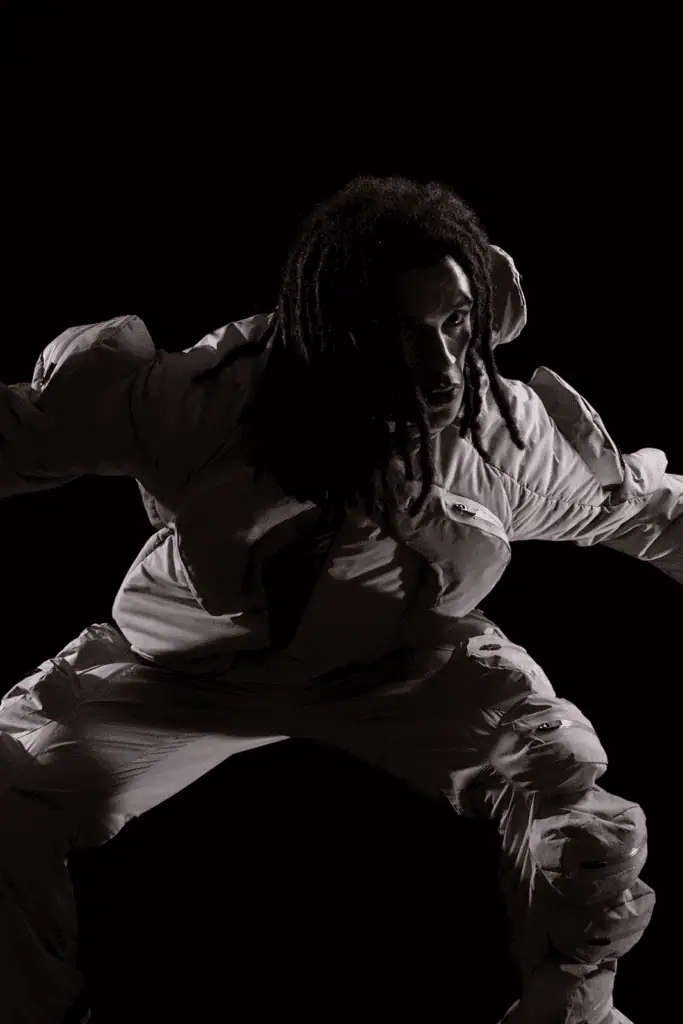 A person with long, dark hair wearing a light-colored, hooded padded suit poses against a dark background, partially crouched with arms outstretched. Dramatic lighting highlights the contours of their face and outfit.
