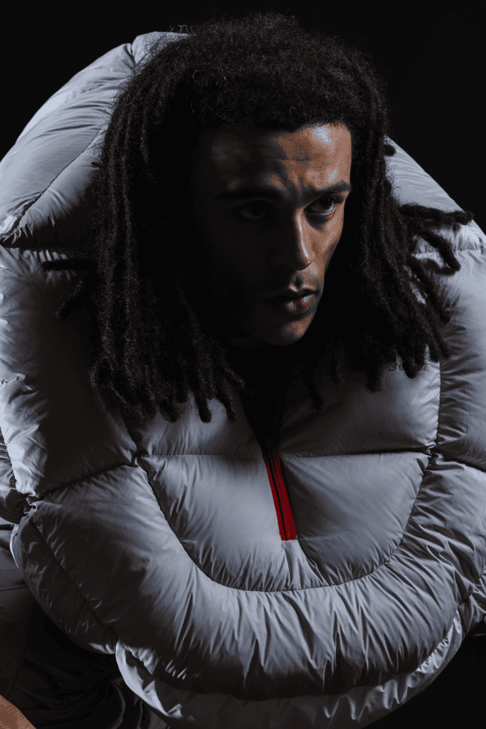 A person with long, dark dreadlocks wears a large, light gray hooded puffer jacket with a prominent red zipper, set against a dark background. Their expression is focused and intense.