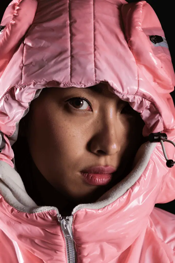 A person with smooth skin and neutral makeup looks directly at the camera, wearing a hooded, shiny pink jacket with white fleece lining, partially zipped up to their chin.