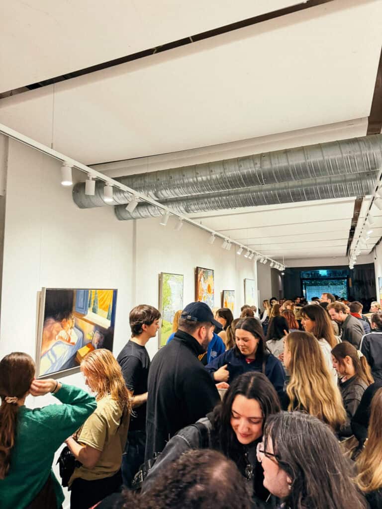 A large crowd of people gathers in a brightly lit art gallery, viewing paintings on the white walls while chatting and mingling. Exposed ductwork is visible along the ceiling.