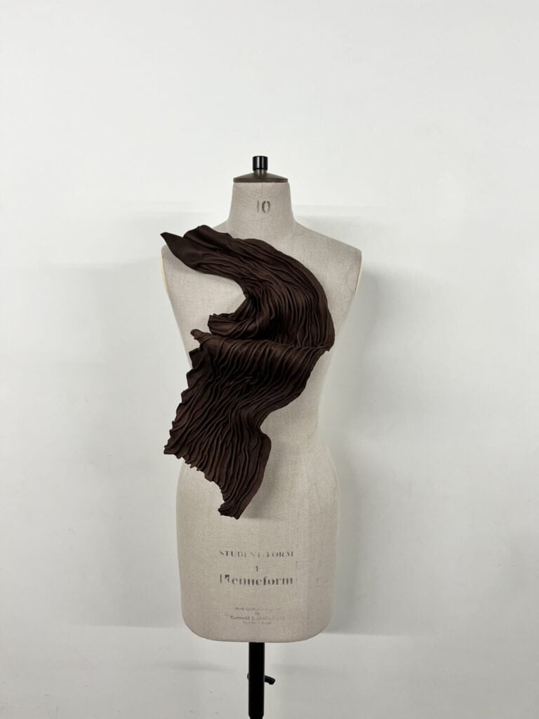 A dress form displays a sculptural, asymmetrical brown garment with dramatic pleats cascading diagonally across the torso against a plain white background.