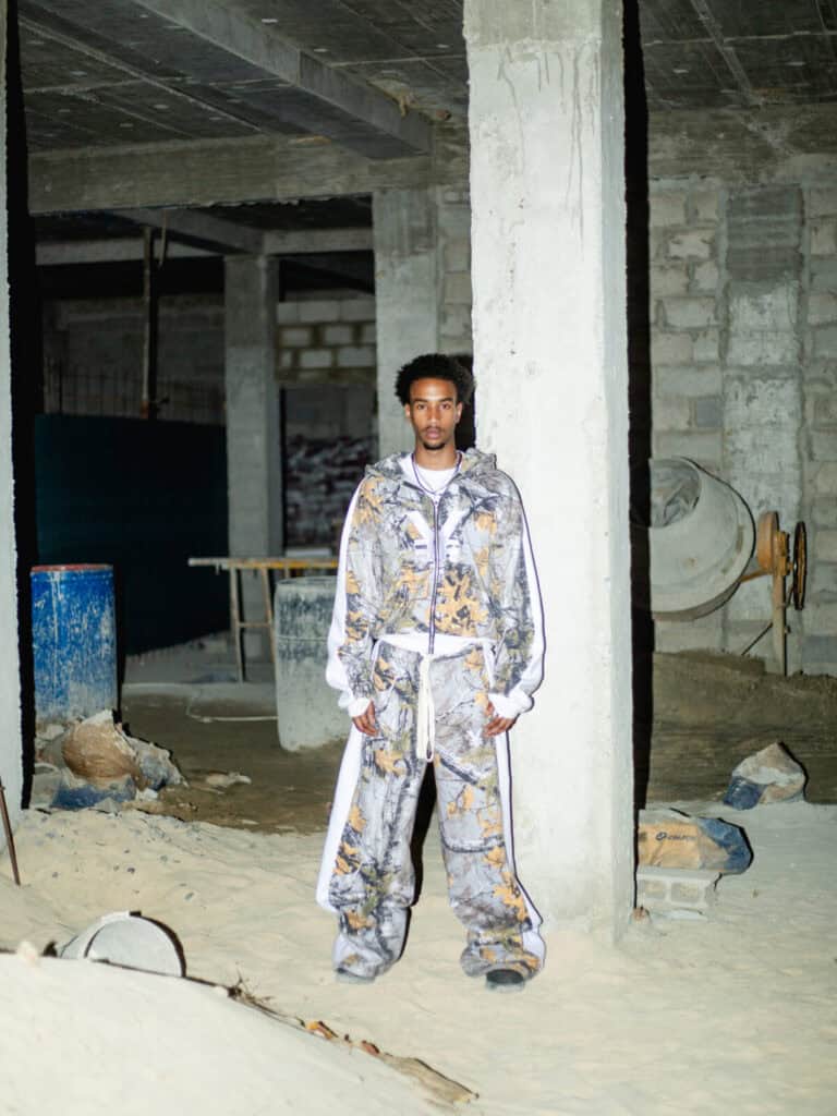 A man wearing a matching patterned hoodie and baggy pants stands in a dimly lit, unfinished concrete building with sand and construction materials scattered around.