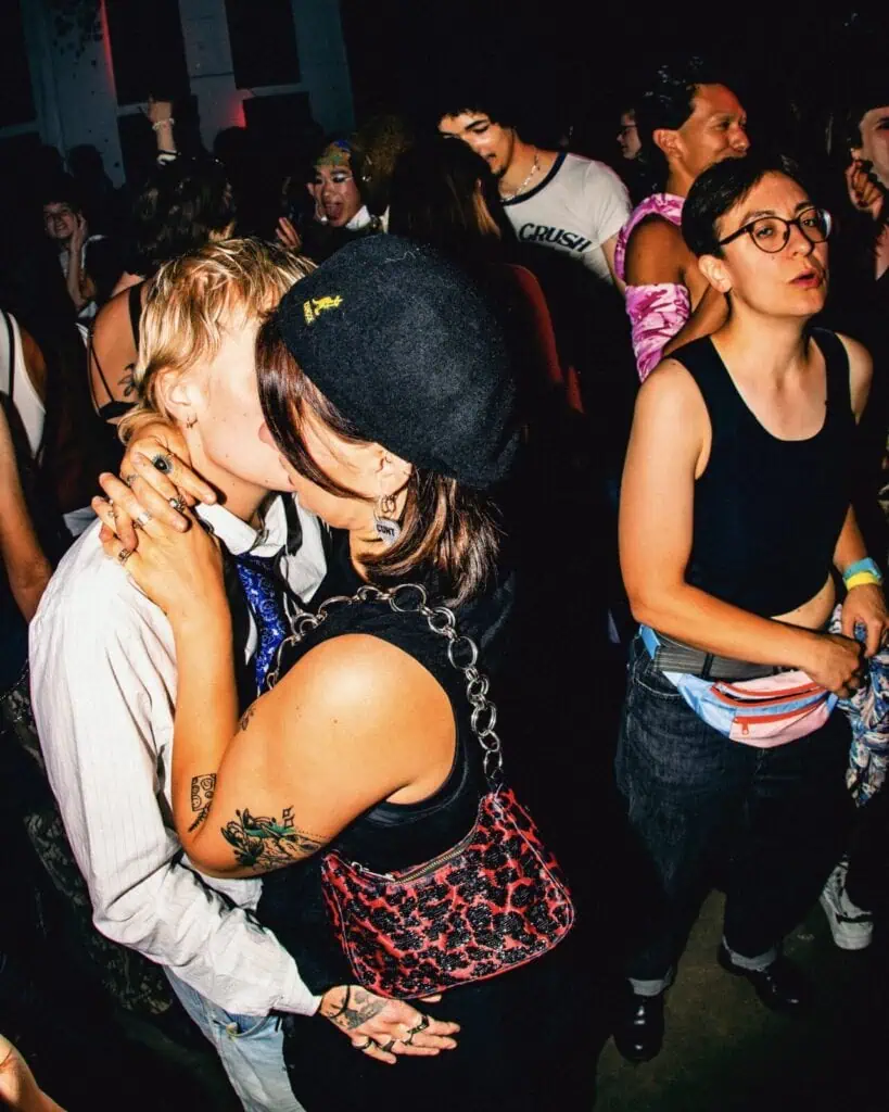 Two people kiss passionately in a crowded, colorful party setting. Other partygoers stand around them, some smiling and talking. The atmosphere is lively and energetic, with bright lighting and vibrant outfits.