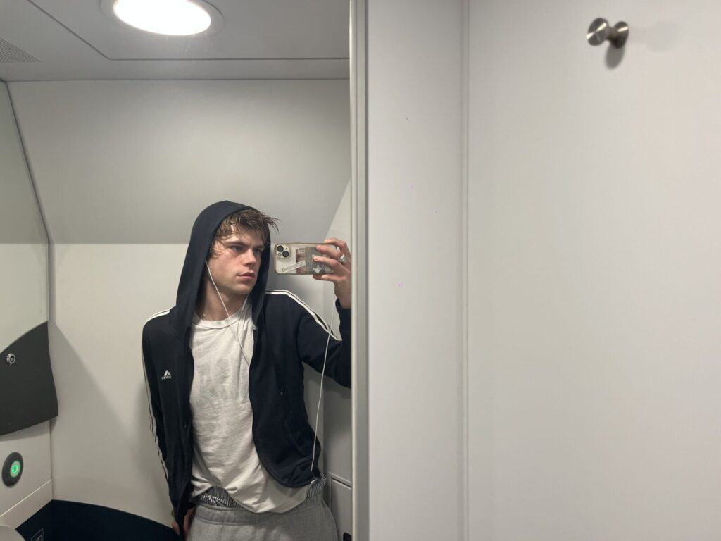 A young person wearing a black hoodie, white t-shirt, and gray pants takes a mirror selfie in what appears to be an airplane bathroom. They are holding a phone and looking slightly to the side.