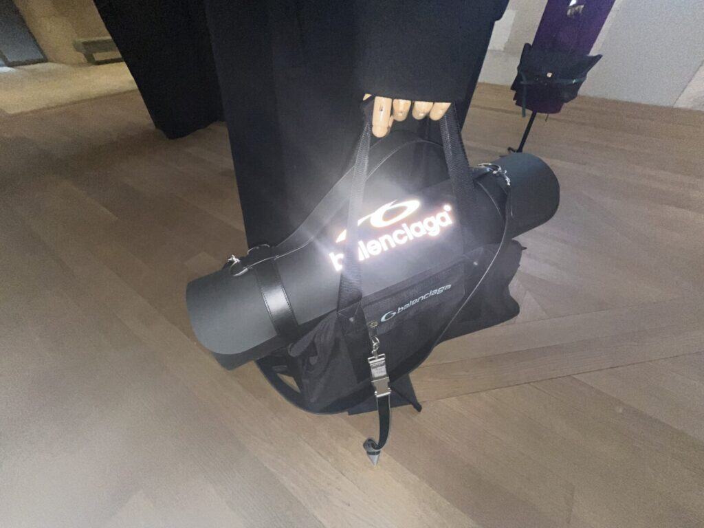 A person wearing black holds a Balenciaga gym bag with a rolled-up yoga mat attached. The Balenciaga logo is illuminated on the side of the bag. The scene is on a light wooden floor.