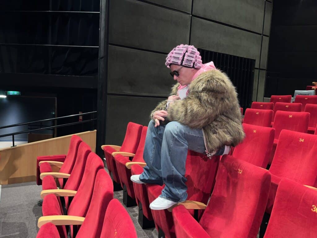 A person wearing a furry jacket, pink knit hat, sunglasses, and jeans sits crouched on a red theater seat in an empty auditorium, looking down thoughtfully.