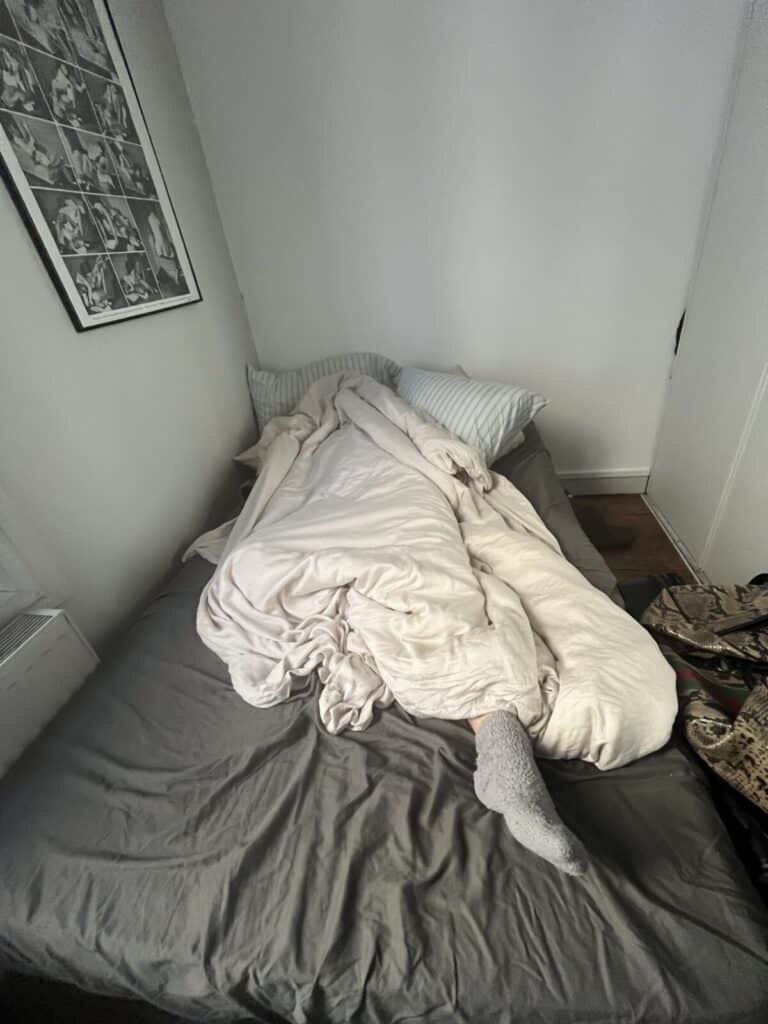 A person is lying in bed mostly covered by a white comforter, with one leg and a foot in a gray sock sticking out. The bed has gray sheets and striped pillows. A collage of black-and-white photos hangs on the wall above.