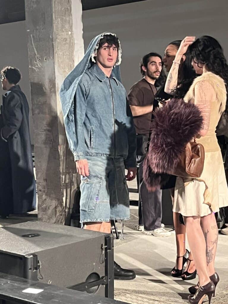 A person wearing an oversized denim hoodie and matching long shorts stands indoors, surrounded by others in fashionable outfits, in what appears to be a fashion event or backstage area.