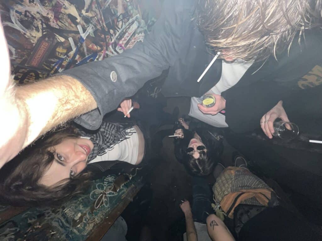 A group of people in a dimly lit, graffiti-covered bar, some holding drinks and cigarettes. One person is taking a selfie from above, others look up at the camera. The atmosphere is casual and crowded.